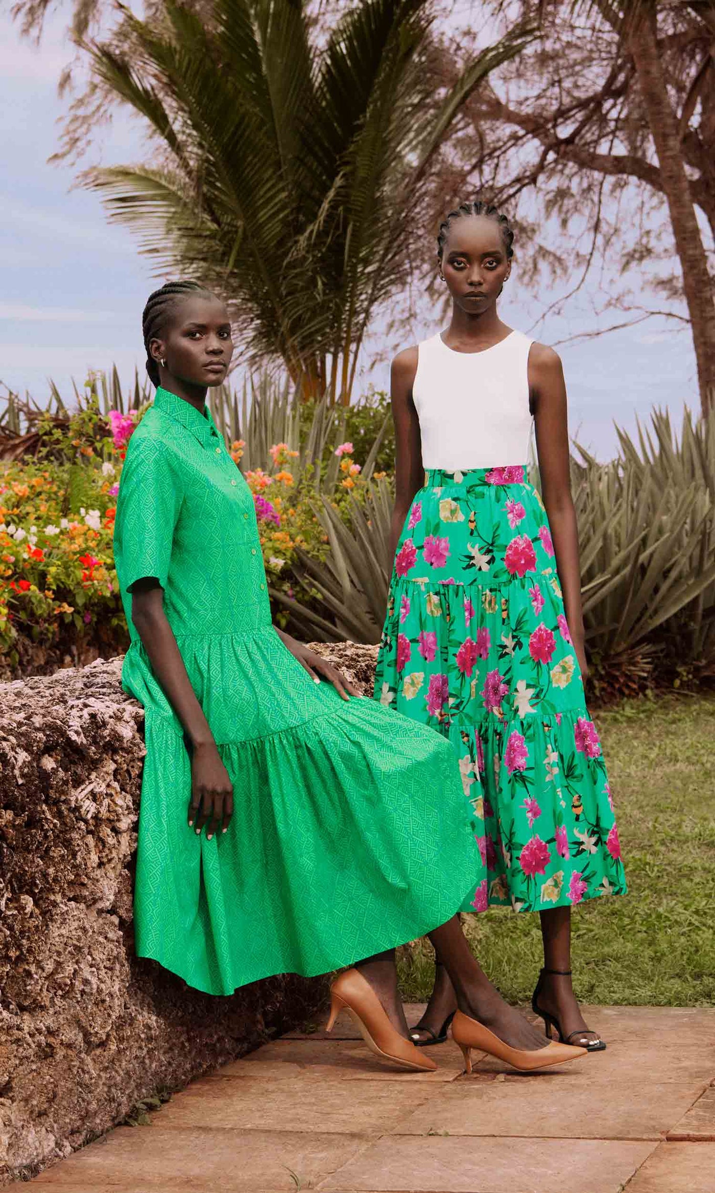 Two models, one sitting on stone wall and the other standing. One wearing a green geometric print tiered dress and the other a cotton poplin tiered skirt in a floral print green